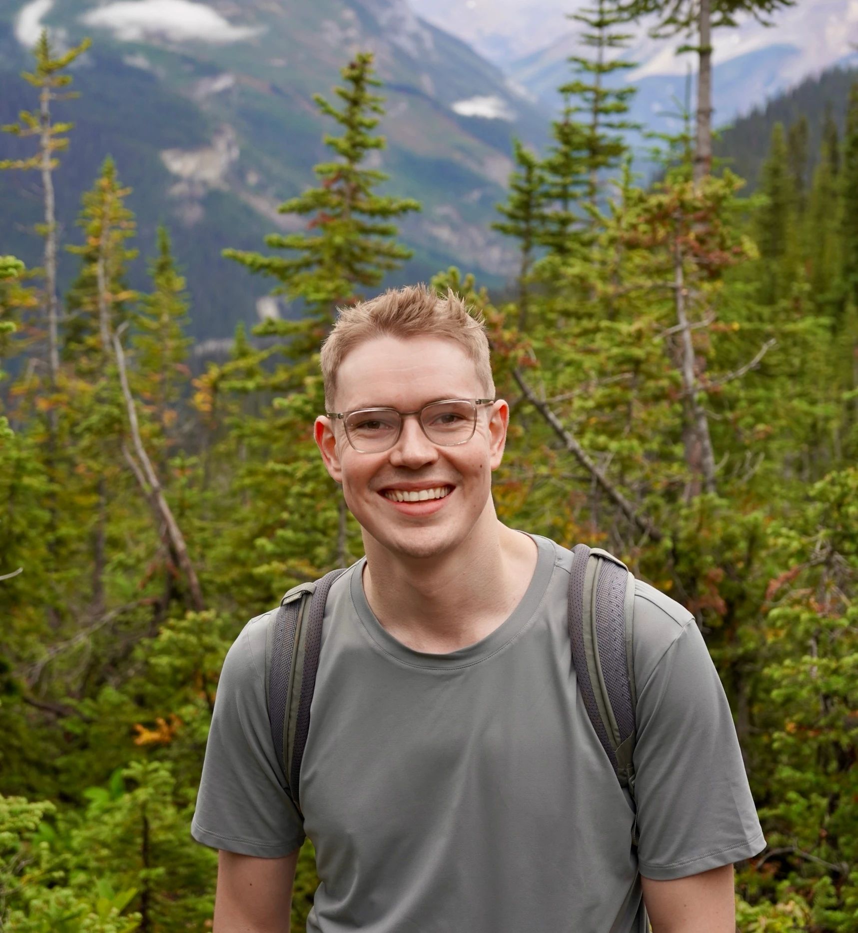 Elliot smiling on a hiking trail, with pine trees and mountains behind him
